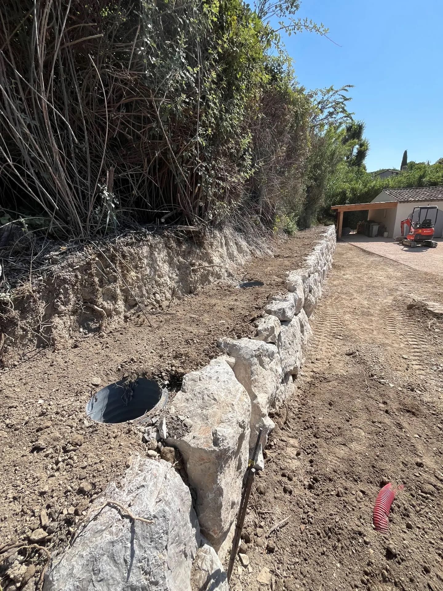 Enrochement de soutènement en cours de montage avec regards de drainage, mini-pelle en arrière-plan