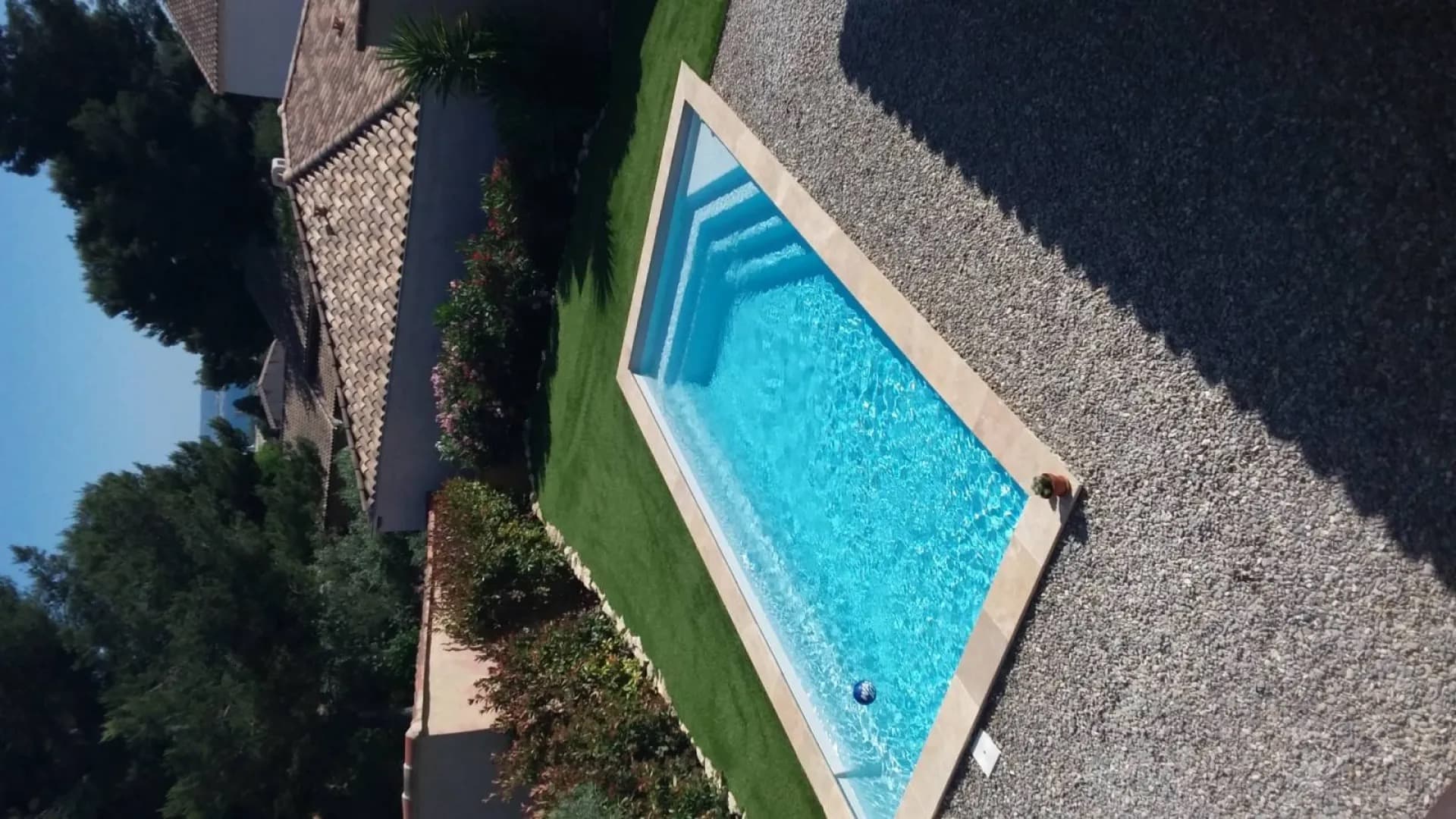 Mini-piscine à margelles claires entourée de gazon synthétique et d’une plage en gravier, le long d’une haie de lauriers-roses
