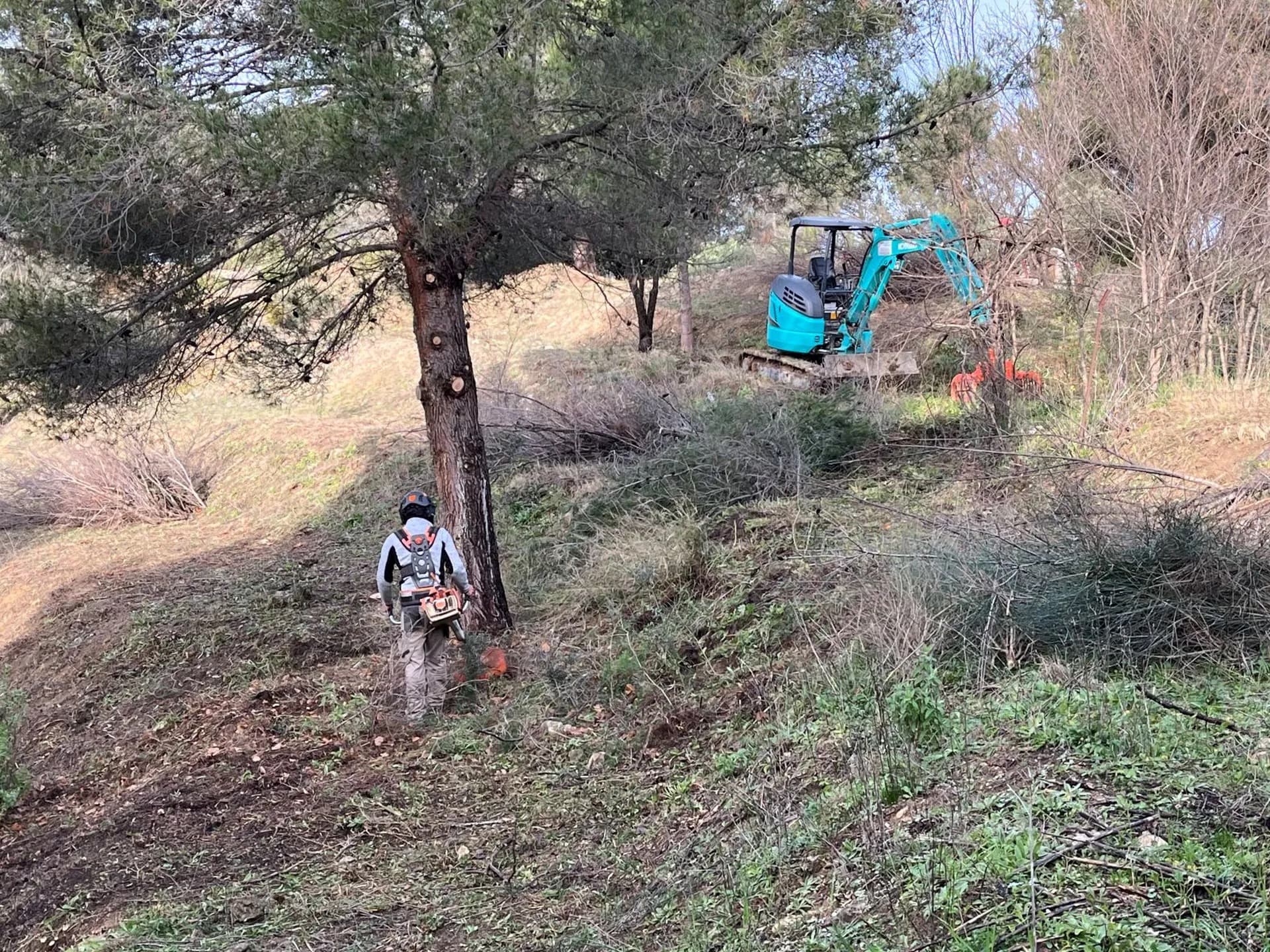 Opérateur AEP casqué, tronçonneuse à la main, et mini-pelle sur un terrain en pente sous les pins à Velaux